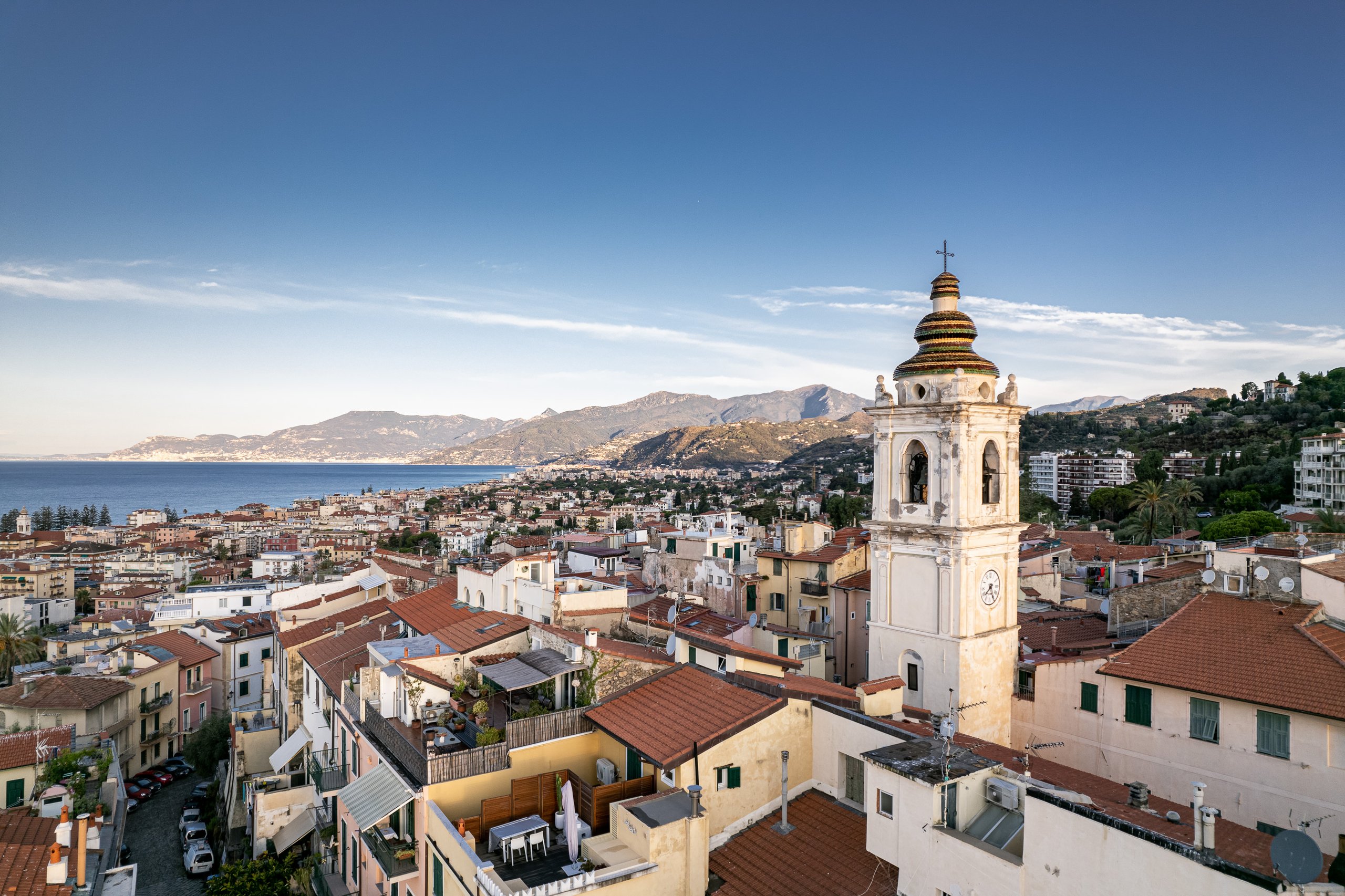 Il campanile bianco di Bordighera Alta sopra i tetti rossi, con la città, il mare e la costa verso la Francia