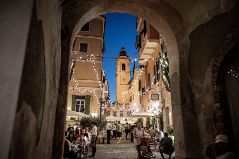 Via di Bordighera Alta con lucine appese, tavoli pieni e il campanile illuminato, vista da un arco di notte