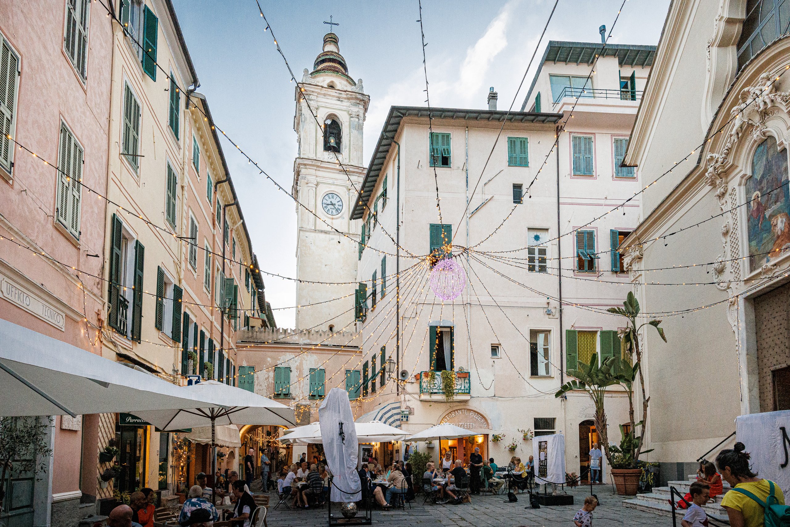 Piazza di Bordighera Alta con il campanile, le luci appese, i dehors affollati e la facciata decorata della chiesa