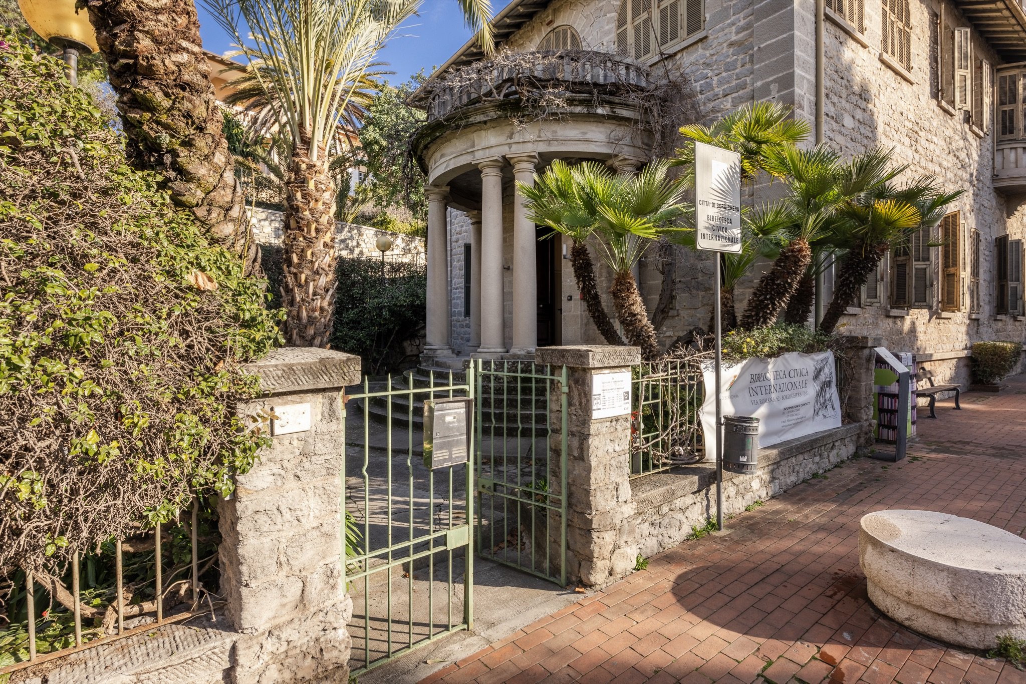 Cancello verde e vialetto in mattoni davanti alla Biblioteca Civica Internazionale, con palme nane e il portico a colonne