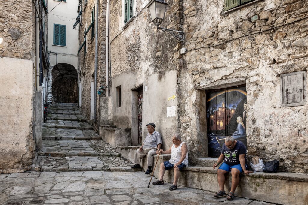Tre anziani seduti su una panca in pietra in un vicolo del Borghetto San Nicolò, accanto a una porta dipinta