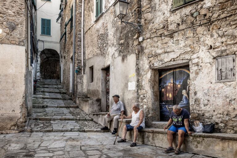 Drei ältere Männer auf einer Steinbank in einer Gasse von Borghetto San Nicolò, neben einer bemalten Tür