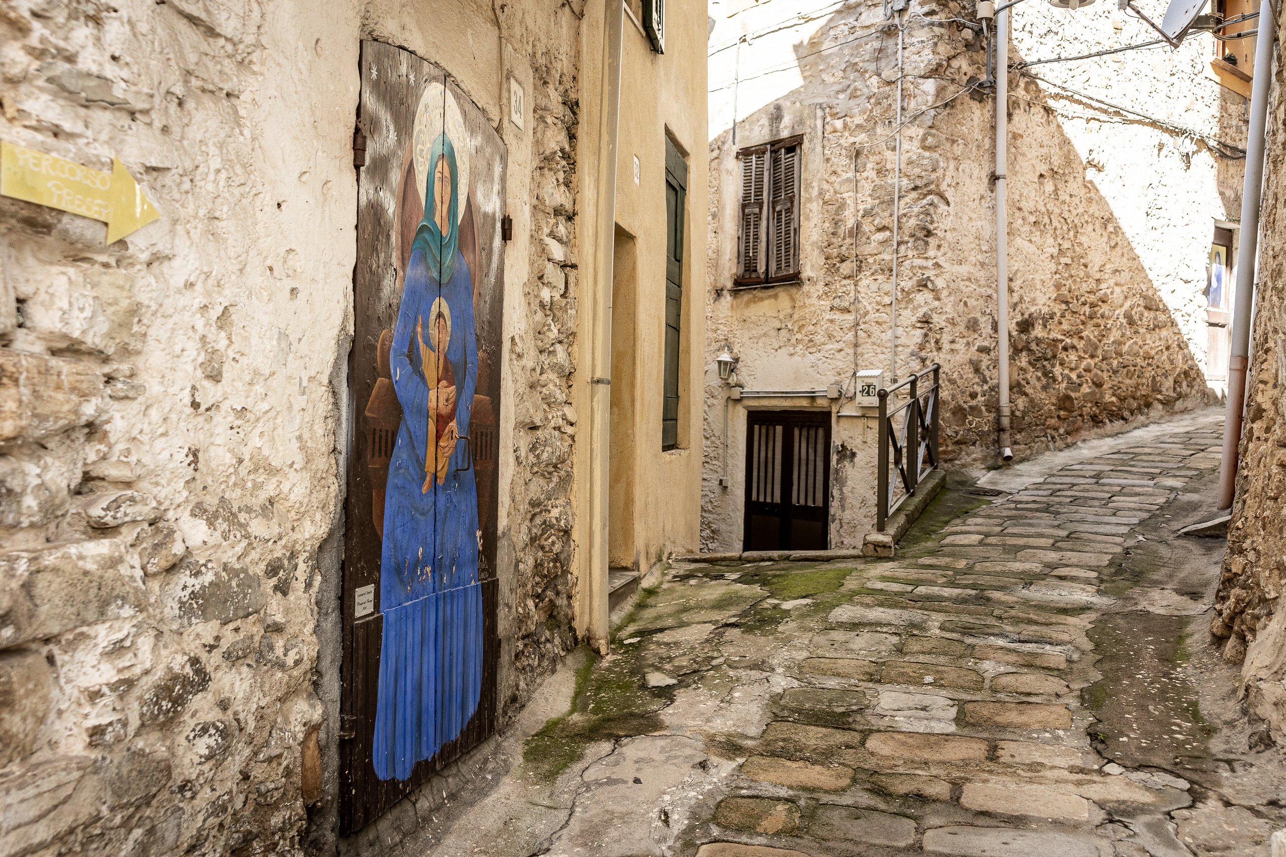 A door painted with a Madonna in a blue cloak holding the Child, in an uphill alley of Borghetto San Nicolò