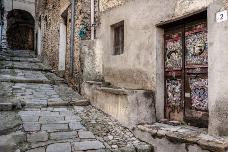 Porta in legno decorata con motivi colorati accanto a una scalinata in pietra in un carruggio del Borghetto San Nicolò