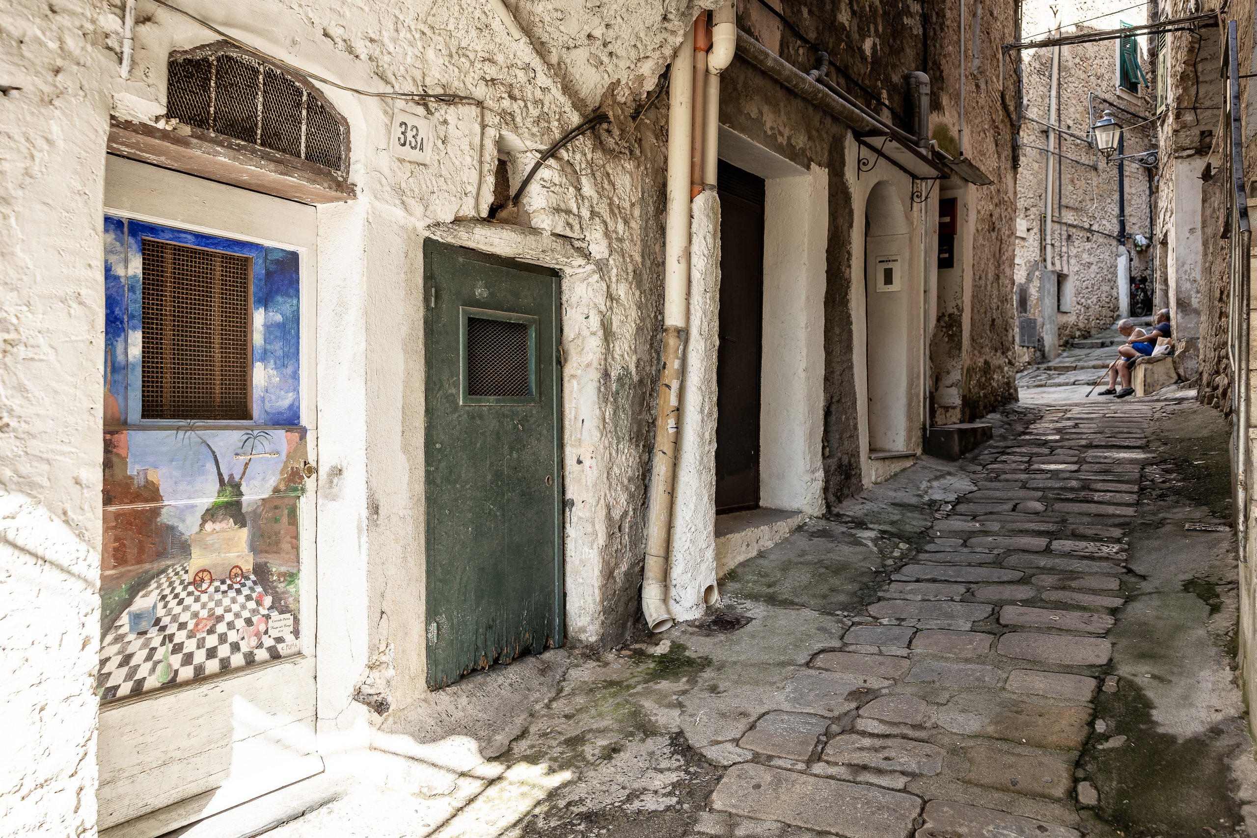 A door painted with a surreal landscape and a chequered floor in a stone alley of Borghetto San Nicolò