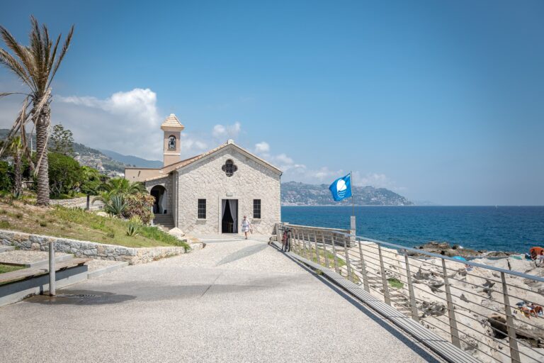 Facciata in pietra della chiesa di Sant'Ampelio con il campanile, la passeggiata e la Bandiera Blu sul mare