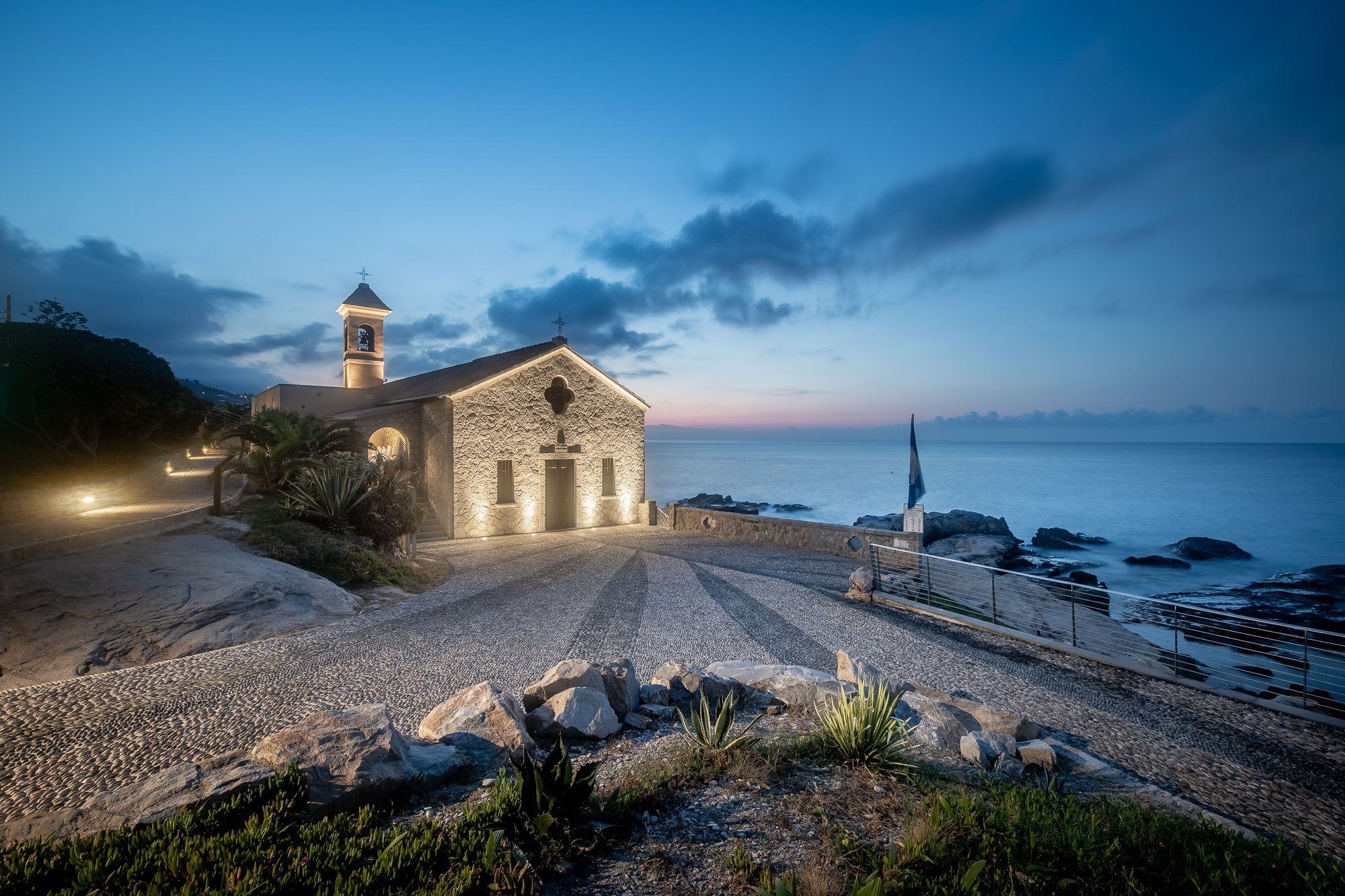 Facciata in pietra della chiesa di Sant'Ampelio illuminata, con il sagrato in ciottoli e il mare calmo al tramonto