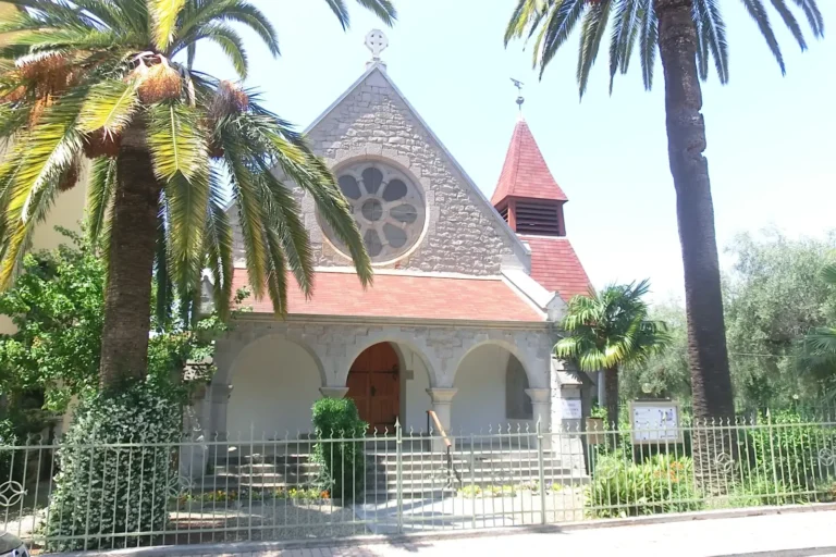 Die Fassade der Waldenserkirche von Bordighera mit Rosette, Bogenportikus und rotem Glockenturm zwischen den Palmen