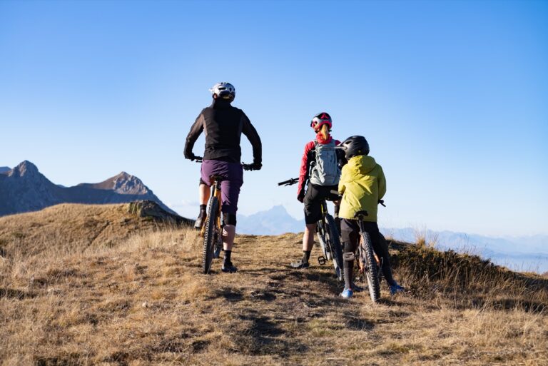 Three riders with mountain bikes on a high-altitude meadow, with the peaks of the Ligurian Alps on the horizon