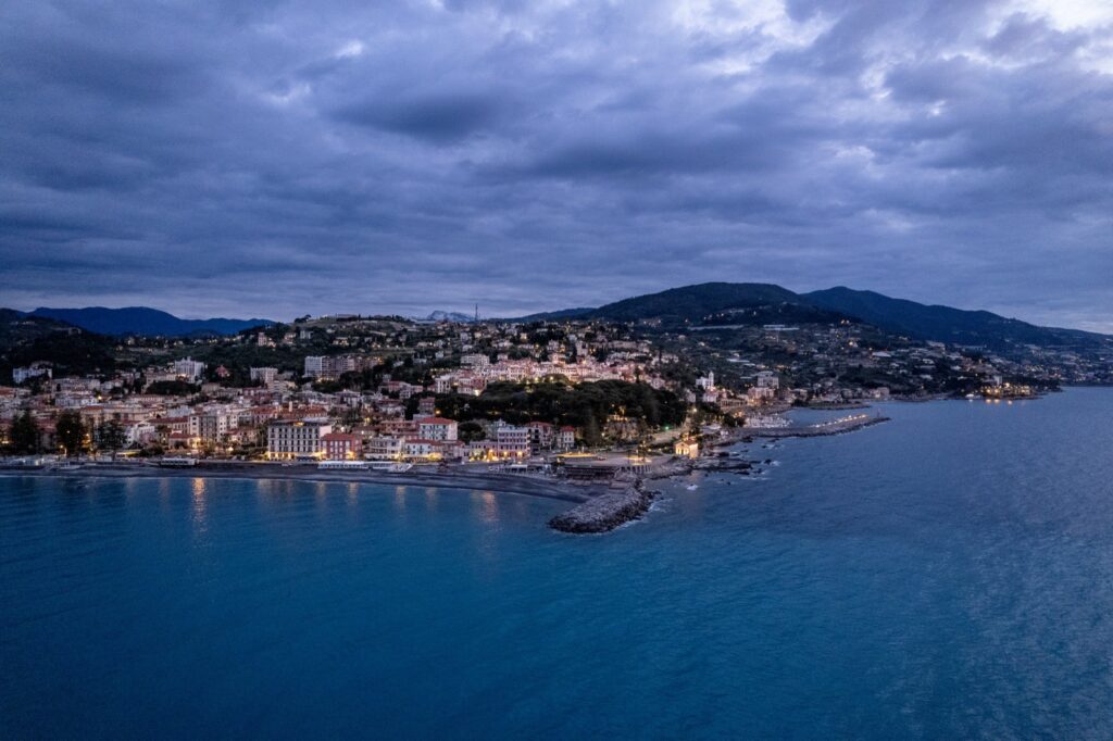 Luftaufnahme von Bordighera zur blauen Stunde, mit der beleuchteten Strandpromenade, dem Hügel und einem wolkenreichen Himmel