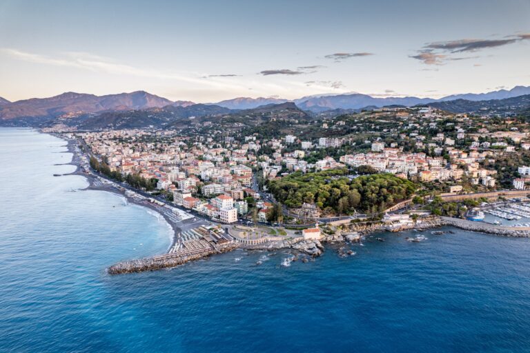 Panorama aereo di Bordighera all'alba, con la città, la pineta del Capo, la spiaggia, il porto e le montagne sullo sfondo