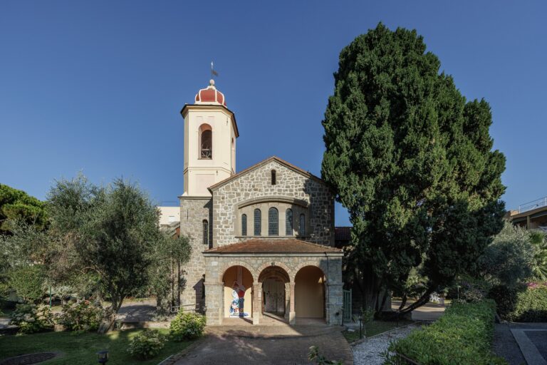 L'ex Chiesa Anglicana con il portico a tre archi, il campanile con cupola rossa e un grande cipresso, sotto il cielo blu