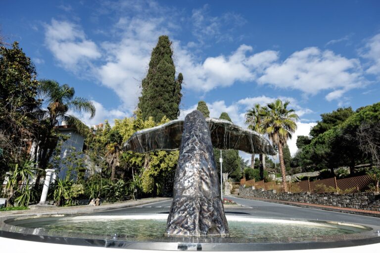 Fontana con la scultura in acciaio di una coda di capodoglio che esce dall'acqua, tra cipressi e palme sotto il cielo azzurro