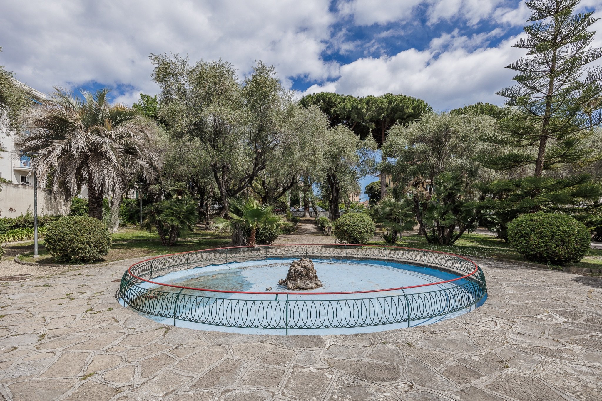 Fontana circolare con ringhiera in ferro al centro dei Giardini Lowe, tra palme, olivi e un'araucaria