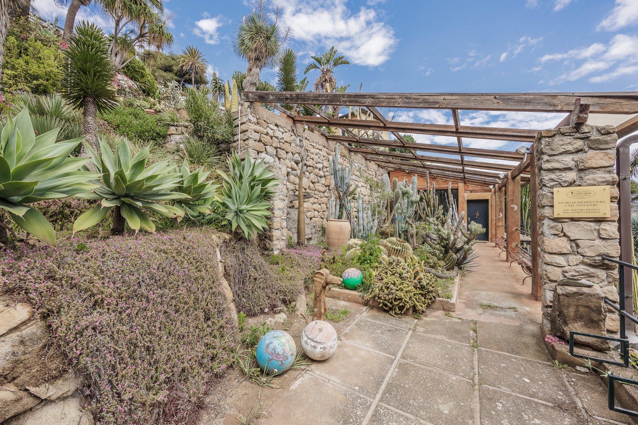 Viale d'ingresso del Giardino Esotico Pallanca con agavi, pergola in legno, muri in pietra e palme