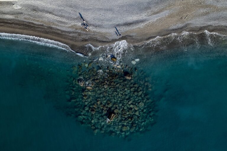 Blick von oben auf einen Wellenbrecher aus Felsblöcken im türkisfarbenen Meer neben einem Kiesstrand an der Blumenriviera