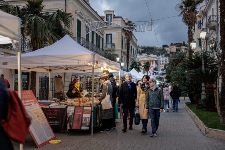 Famiglia a passeggio tra le bancarelle illuminate del mercatino di Natale nel centro di Bordighera, tra palme e palazzi