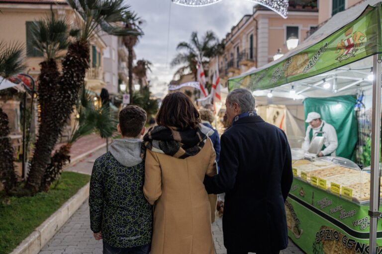Famiglia a passeggio tra le bancarelle illuminate del mercatino di Natale nel centro di Bordighera, tra palme e palazzi