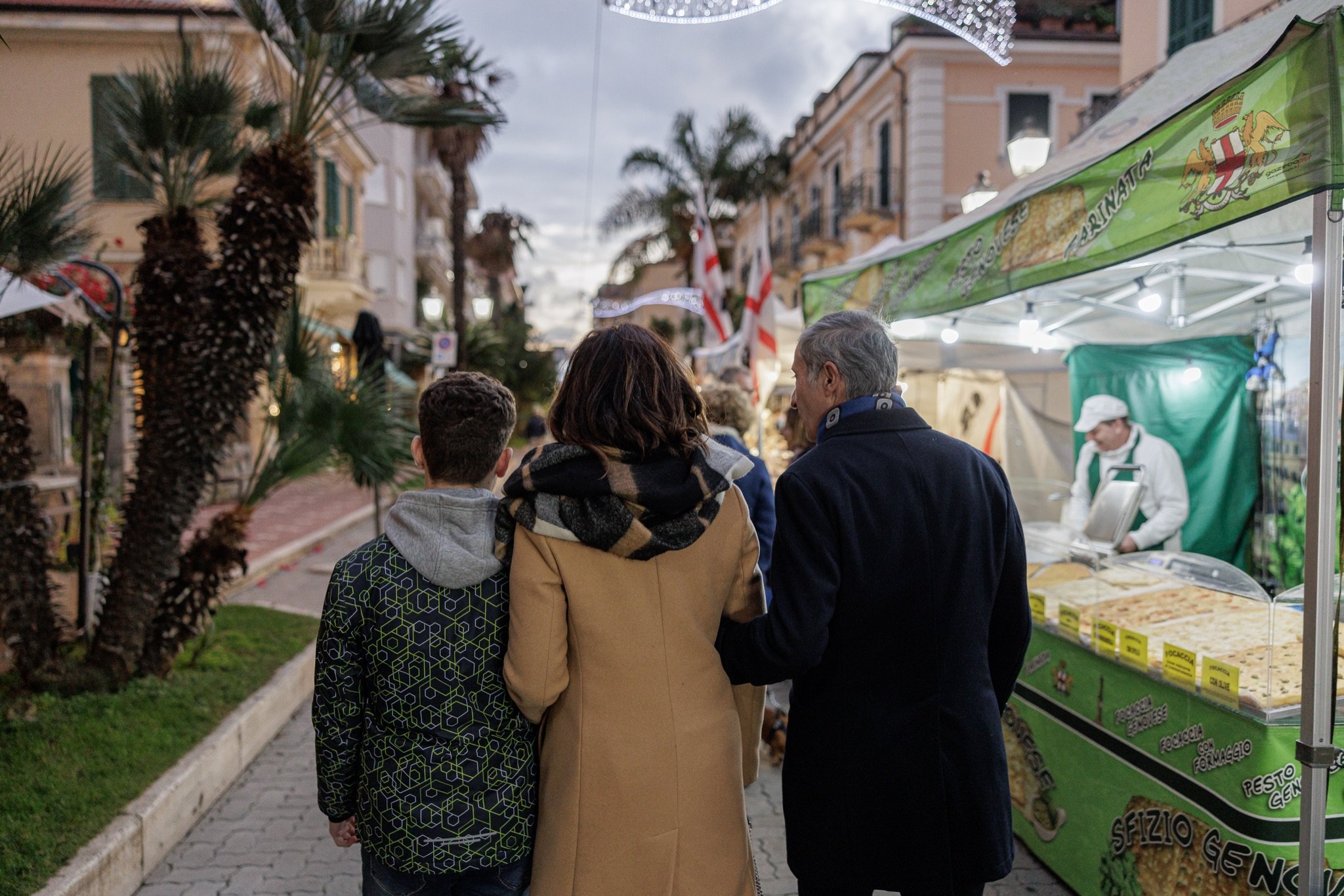Famiglia a passeggio tra le bancarelle illuminate del mercatino di Natale nel centro di Bordighera, tra palme e palazzi