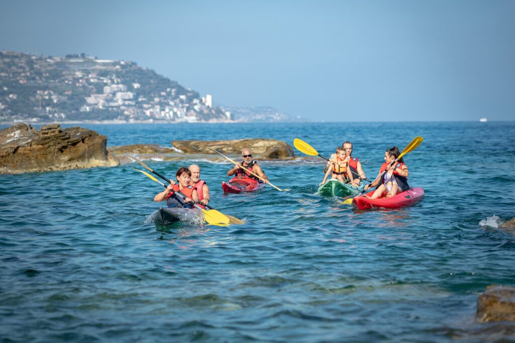Gruppo in kayak rossi, verdi e neri che pagaia vicino agli scogli, con il promontorio e la costa di ponente sullo sfondo