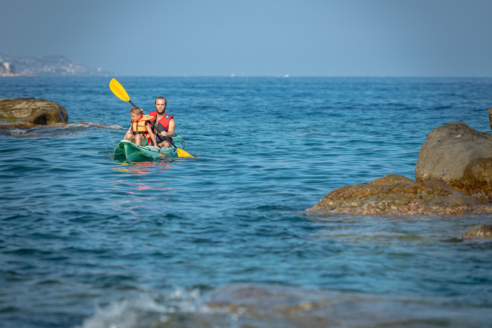 Kayak verde con due persone in giubbotto salvagente davanti a scogli bruni, nel mare azzurro di Bordighera