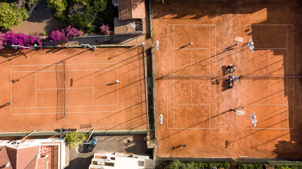 Vista aerea di due campi in terra rossa del Bordighera Lawn Tennis Club con giocatori, ombrelloni e bouganville fucsia