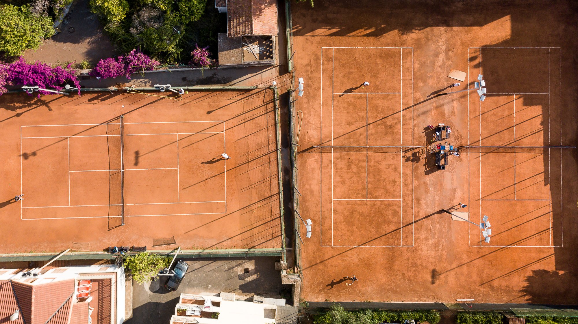 Vista aerea di due campi in terra rossa del Bordighera Lawn Tennis Club con giocatori, ombrelloni e bouganville fucsia