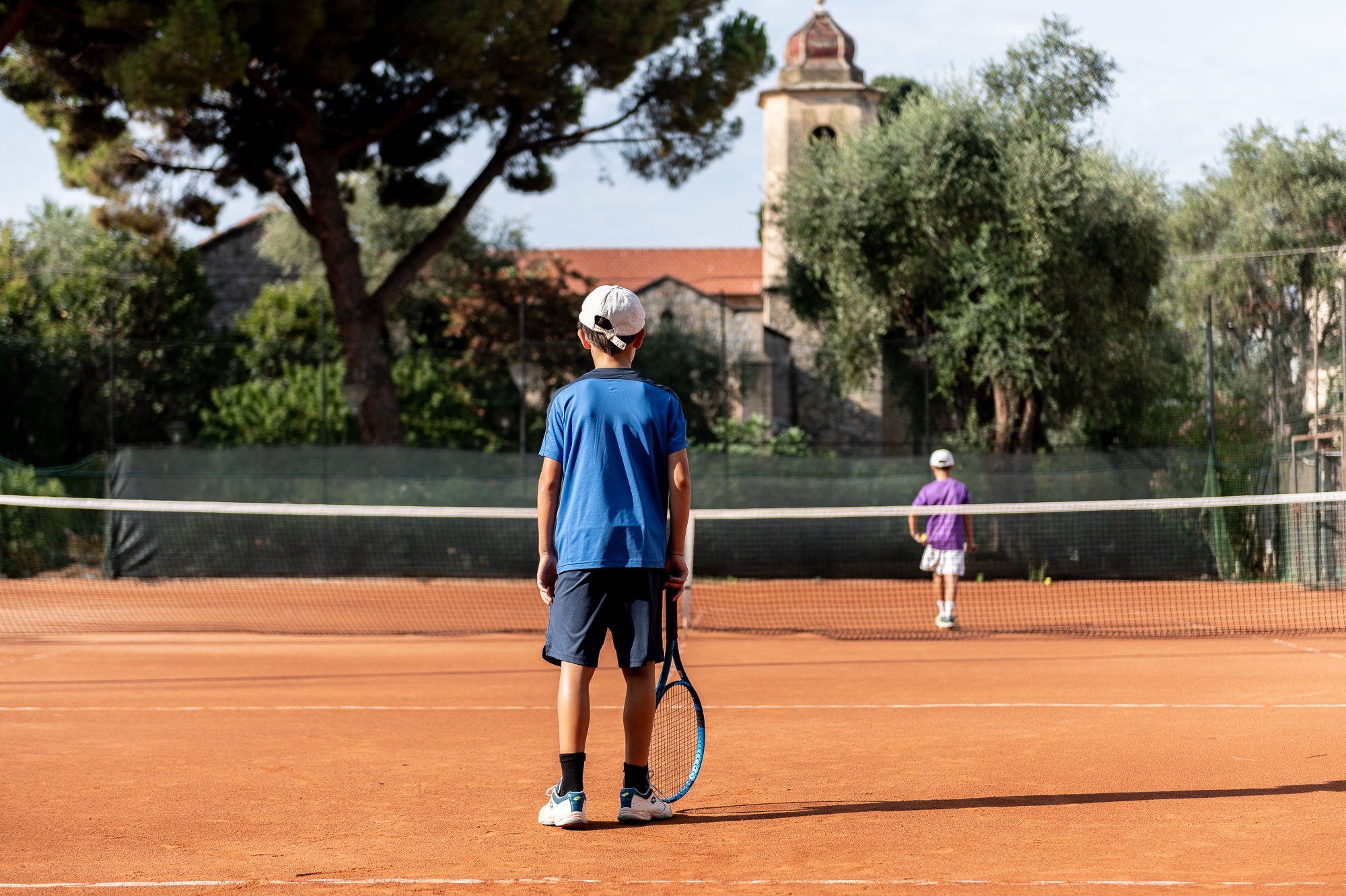 Due giovani tennisti ai lati della rete su un campo in terra rossa, con pini, olivi e il campanile dell'ex Chiesa Anglicana