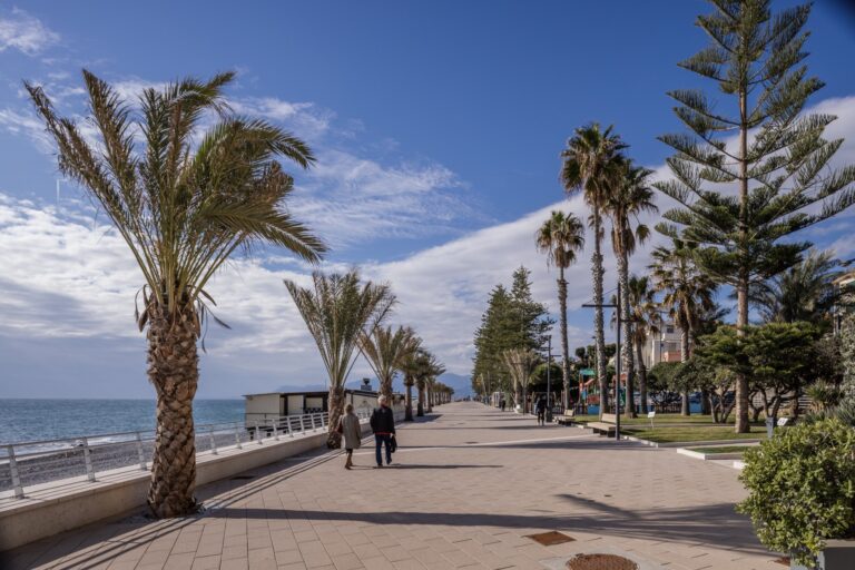 Passanti sulla passeggiata del nuovo lungomare tra palme, araucarie e ringhiera bianca, con il mare e le nuvole