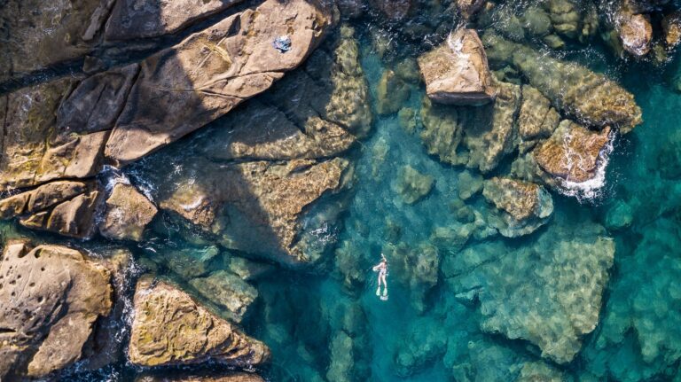 Persona che nuota con maschera tra grandi scogli di arenaria e fondali verdi, vista dal drone sulla costa di Bordighera