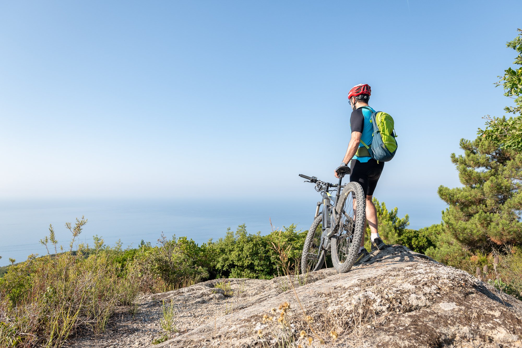 Ciclista in piedi accanto alla mountain bike su una roccia del Monte Nero, con il mare azzurro fino all'orizzonte