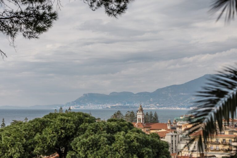 Panorama sui tetti e sul campanile di Bordighera con il mare e la costa francese verso Mentone e Monaco, tra pini e palme