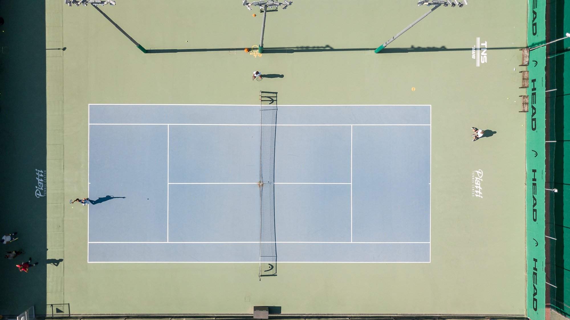 Vista zenitale di un campo in cemento azzurro del Piatti Tennis Center con giocatori e le lunghe ombre