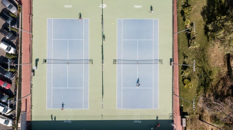 Vista zenitale di due campi in cemento azzurro del Piatti Tennis Center con giocatori, il parcheggio e il verde