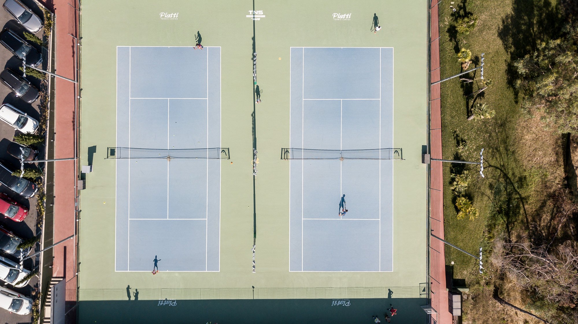 Vista zenitale di due campi in cemento azzurro del Piatti Tennis Center con giocatori, il parcheggio e il verde