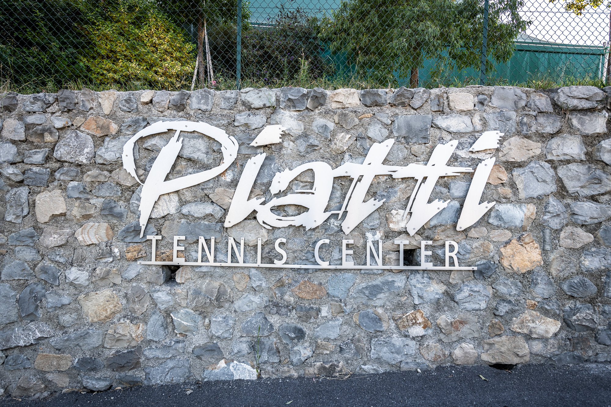 Insegna in metallo Piatti Tennis Center su un muro in pietra, con la recinzione e il verde sopra
