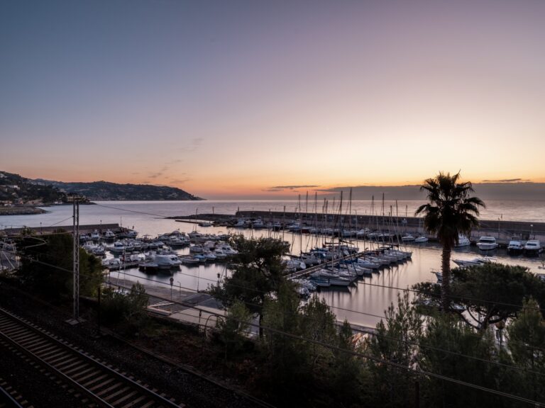 Il porto turistico di Bordighera all'alba con le barche ormeggiate, una palma, la ferrovia e la costa di levante