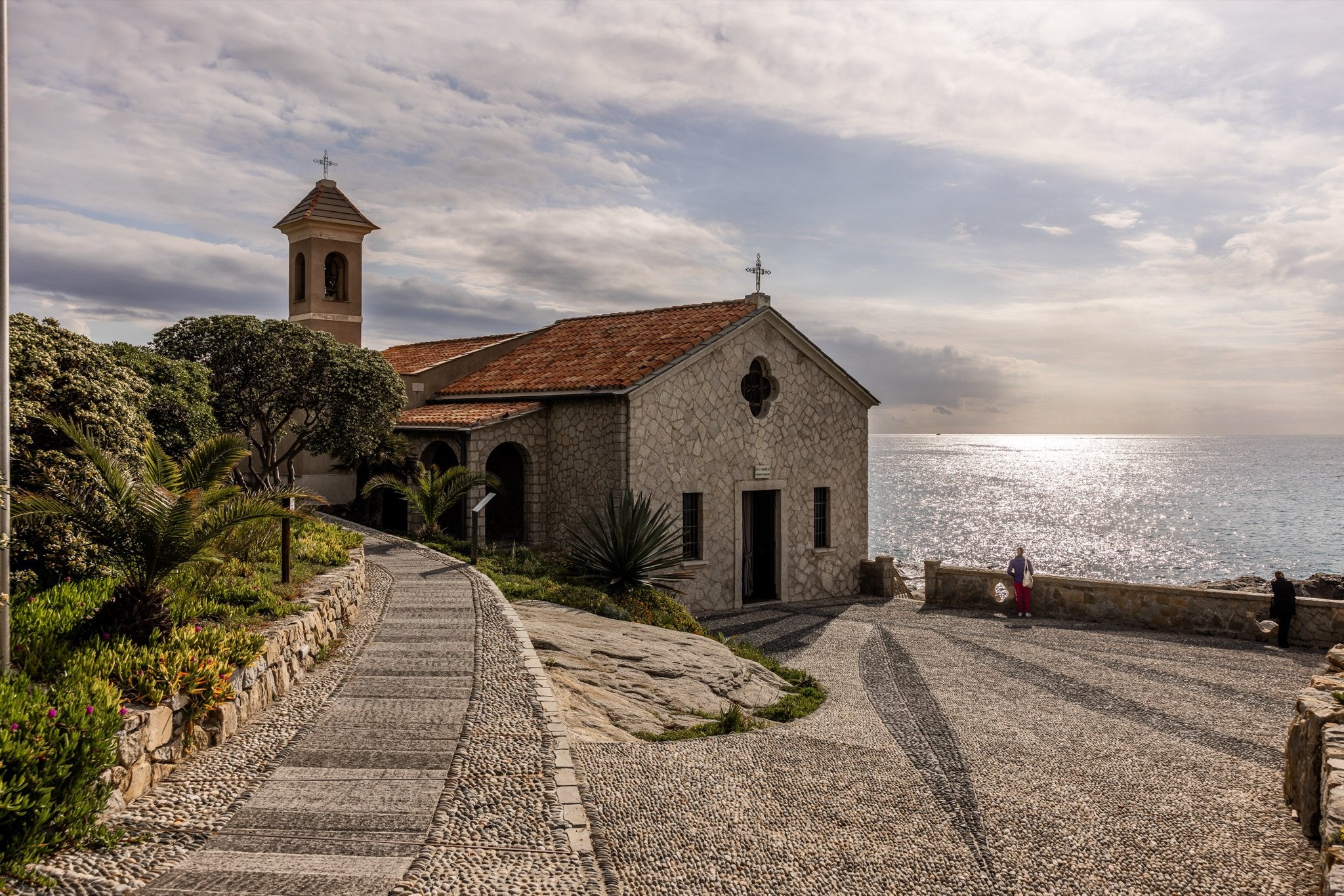 La chiesa di Sant'Ampelio con il campanile, il vialetto e il sagrato in ciottoli, sotto un cielo di nuvole sul mare