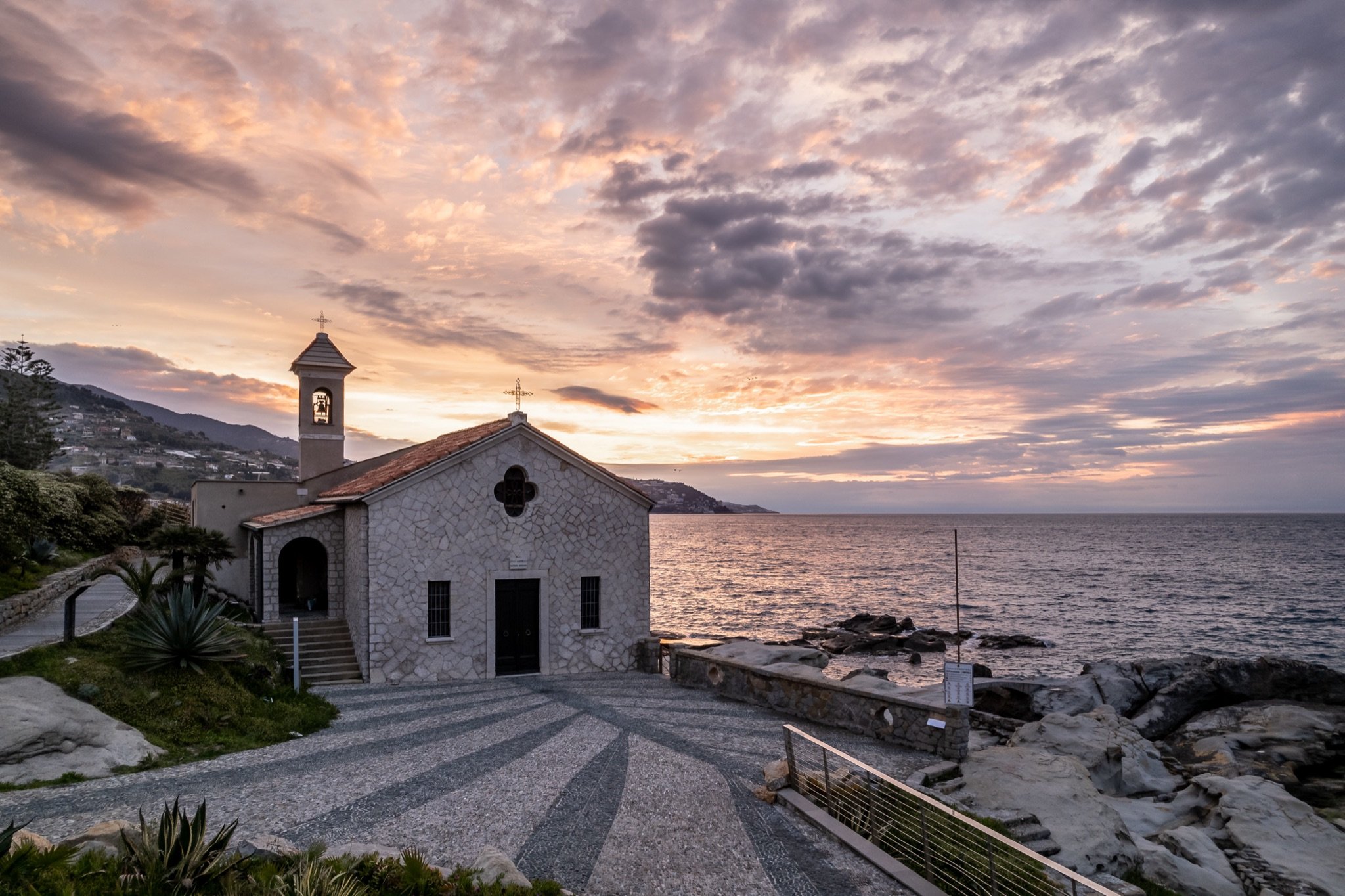 La chiesa di Sant'Ampelio con il sagrato in ciottoli sotto un cielo rosa e arancione al tramonto