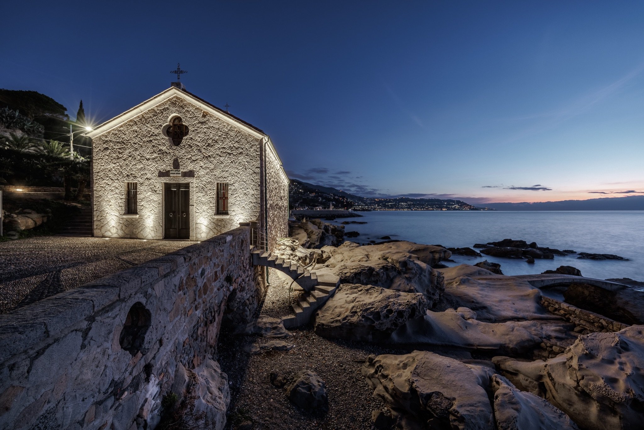 Facciata in pietra della chiesa di Sant'Ampelio illuminata di notte, con gli scogli e la costa sullo sfondo
