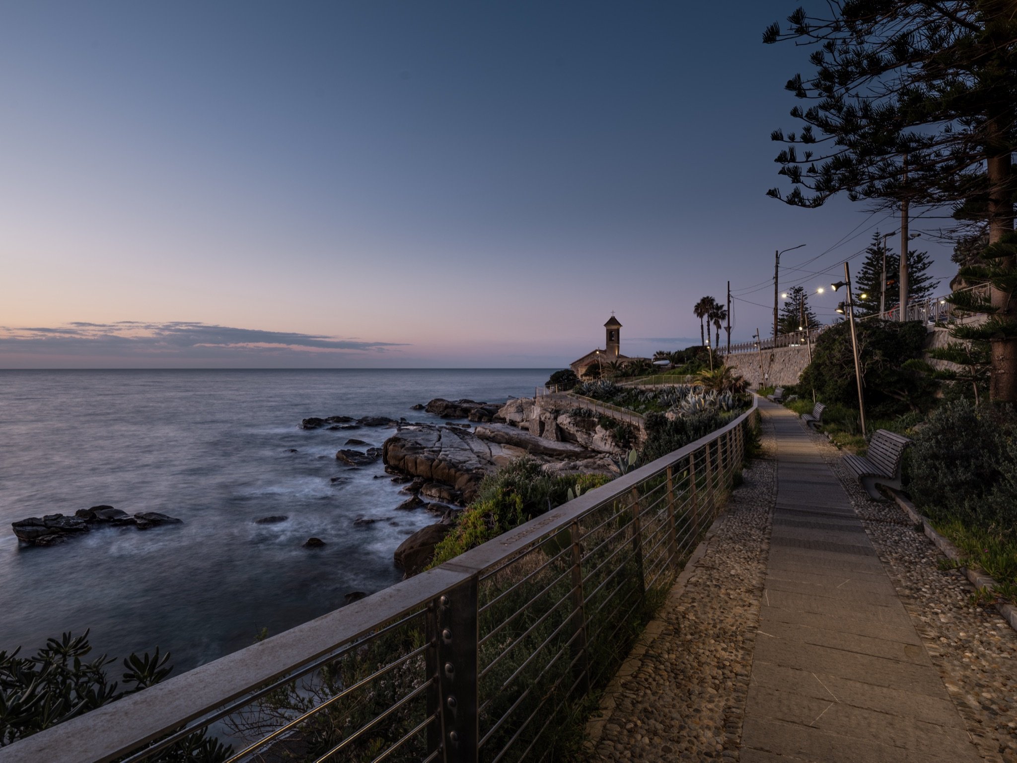 Passeggiata con ringhiera lungo la scogliera verso Sant'Ampelio, con il mare vaporoso all'alba