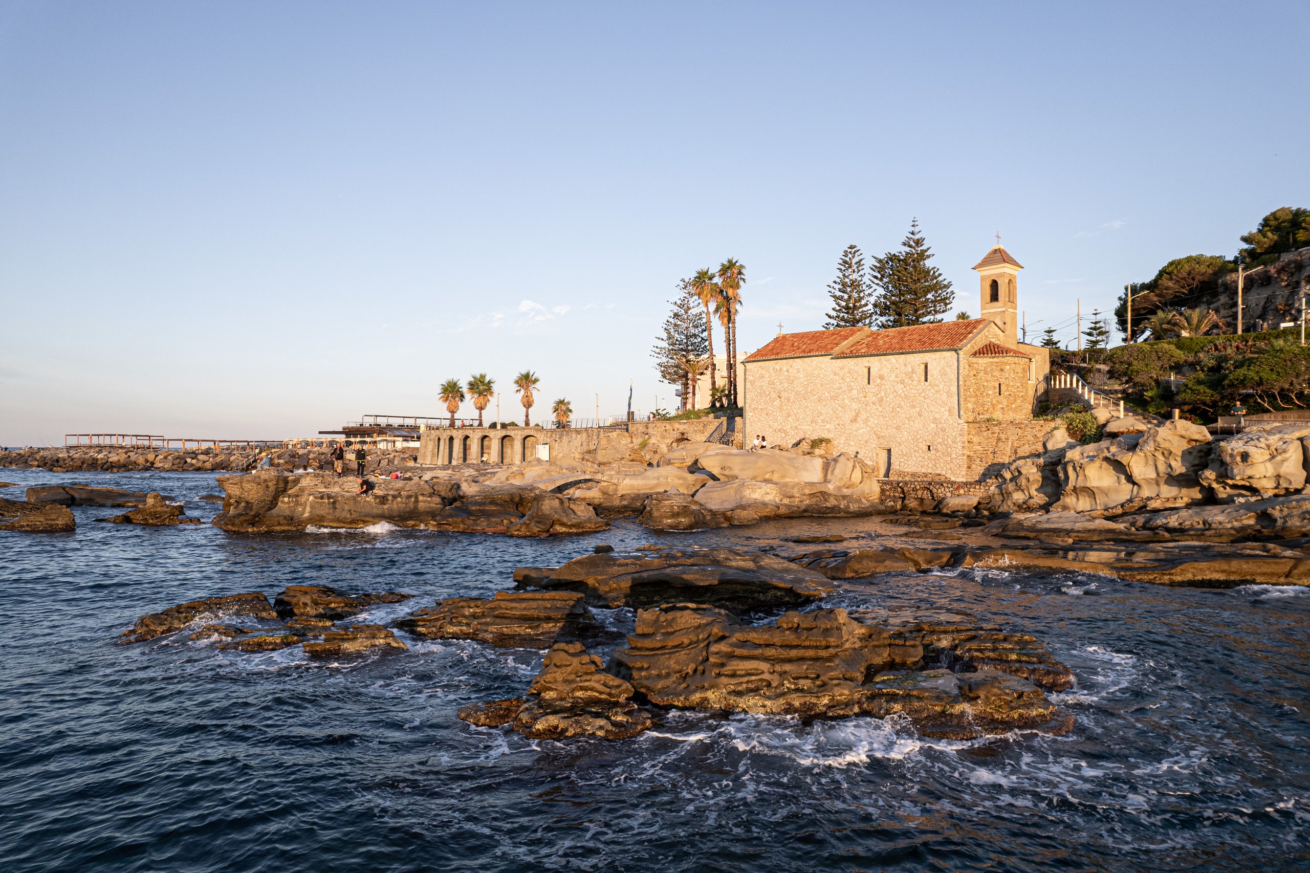Scogli di arenaria stratificata nel mare di Capo Sant'Ampelio, con la chiesetta, i pini e le palme sullo sfondo