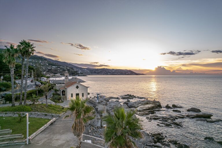 Il sole che tramonta sul mare davanti a Capo Sant'Ampelio, con la chiesetta, le palme e la scogliera in controluce
