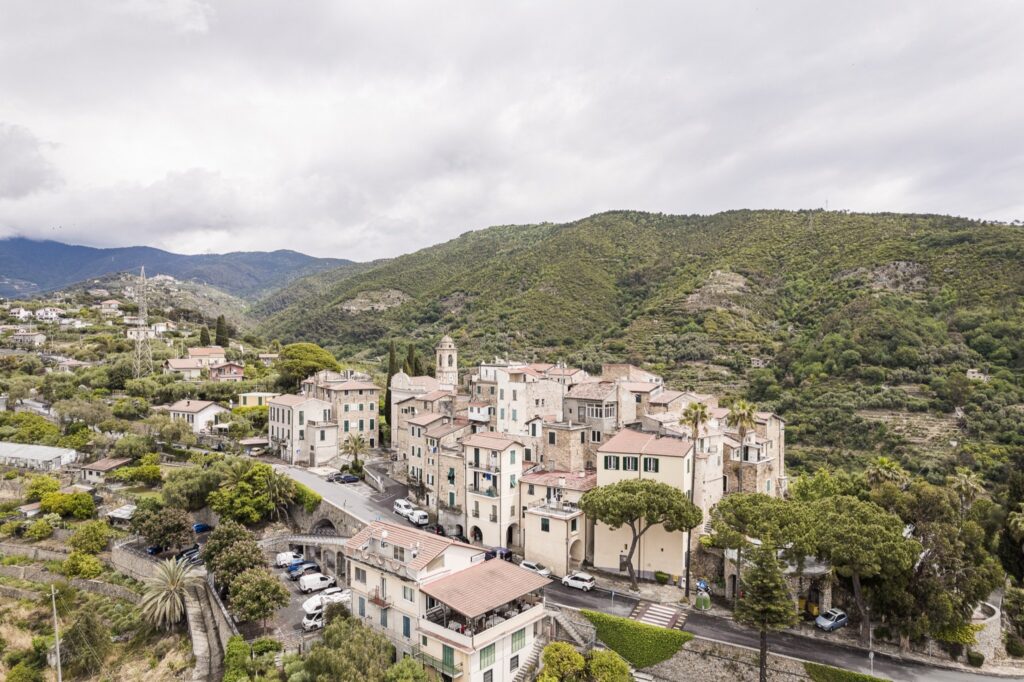 Vista aerea del borgo di Sasso tra colline verdi, olivi e case sparse, con le montagne sullo sfondo