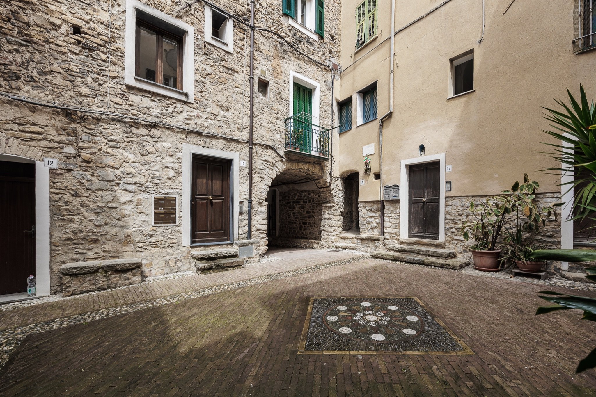 A small brick-paved square with a circular mosaic among stone houses with green shutters and doorways in the village of Sasso
