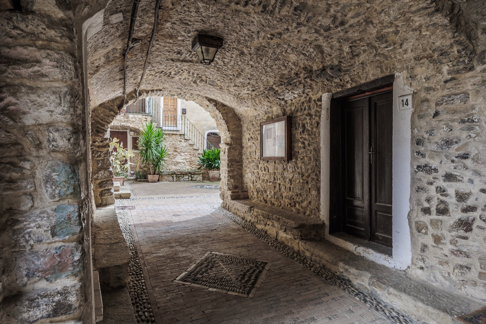 A vaulted passage with a lantern and a wooden door opening onto a small square with plants in the village of Sasso