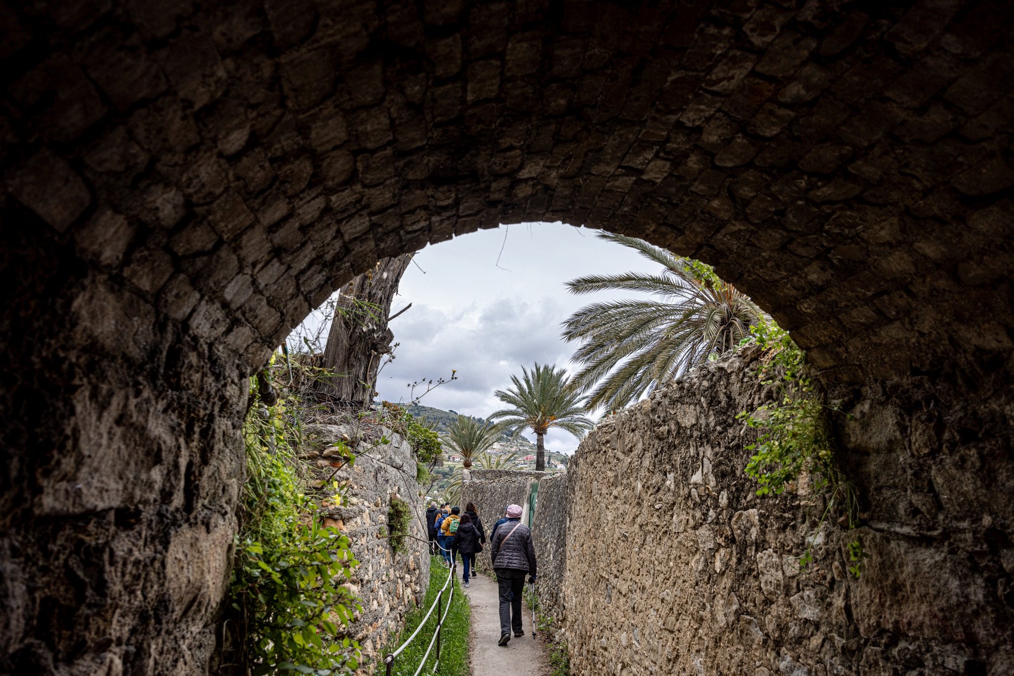 Arco in pietra di una galleria del Beodo che si apre sul sentiero tra muri, palme e un gruppo di escursionisti