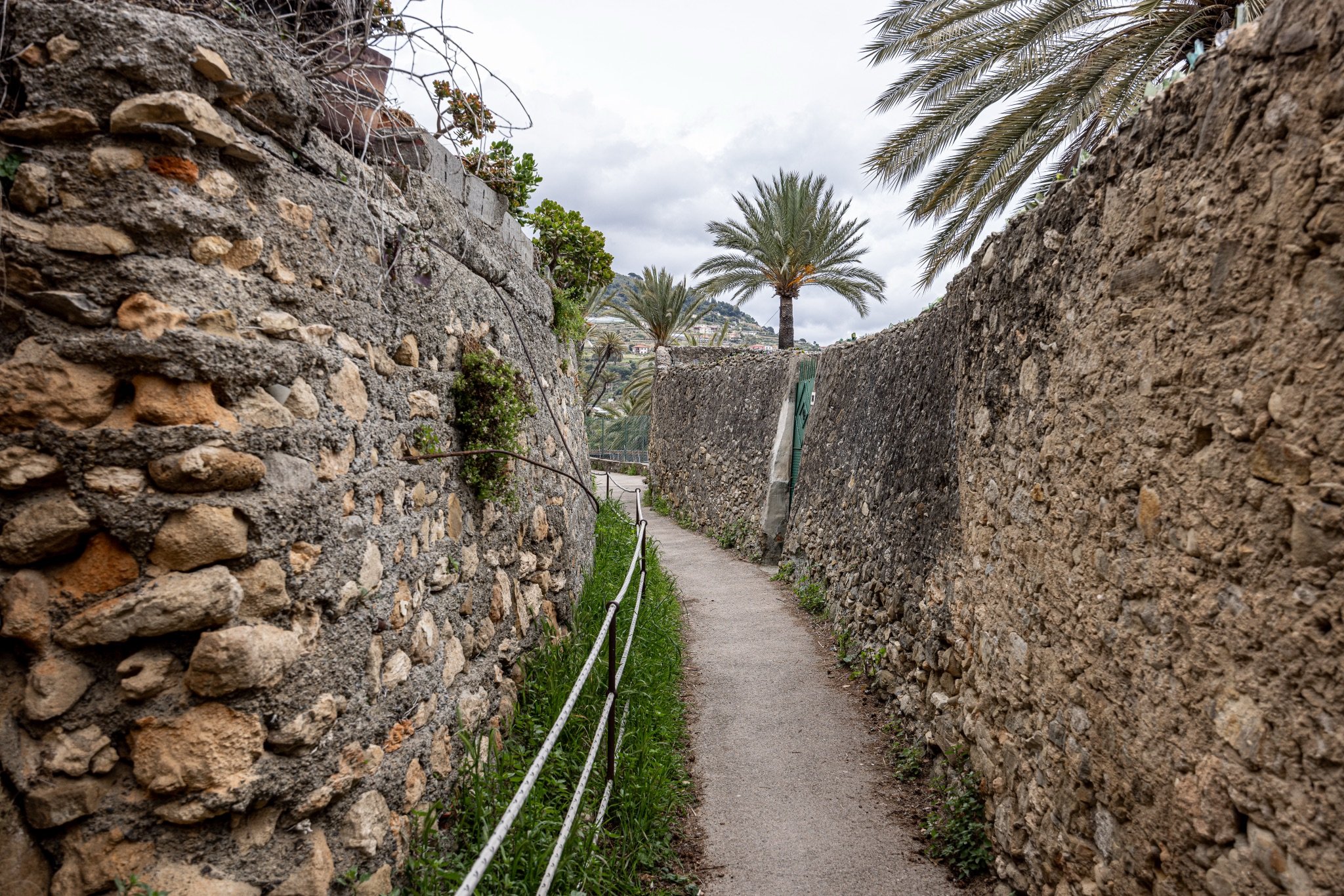 Sentiero del Beodo tra un alto muro in pietra e una ringhiera, con palme da dattero e il cielo nuvoloso