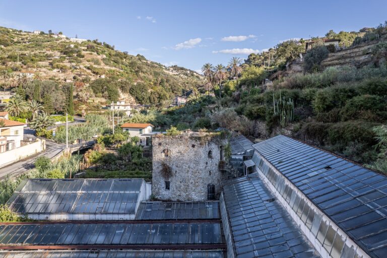 Vecchie serre in vetro e una torre in pietra nel vallone del Sentiero del Beodo, tra palme, fichi d'India e terrazze coltivate