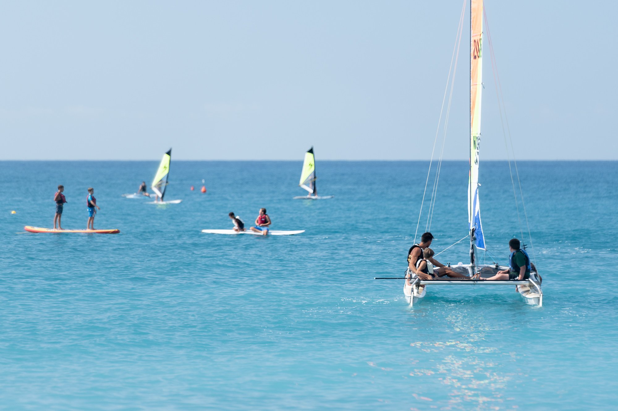 Catamarano con due velisti, persone in SUP e due windsurf nel mare turchese di Bordighera